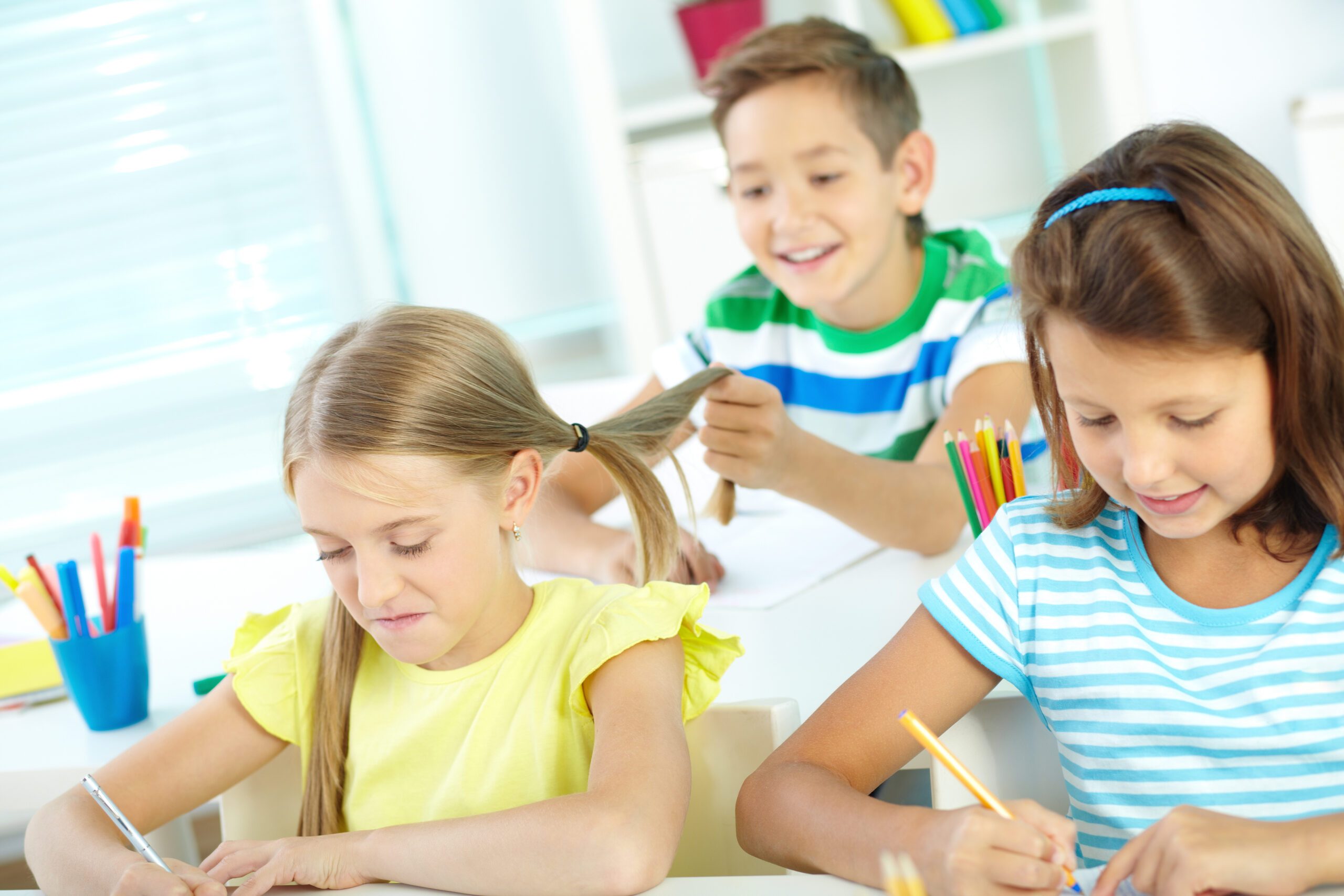 Portrait of lovely schoolgirls drawing at workplace while their schoolmate on background pulling hair of one of them